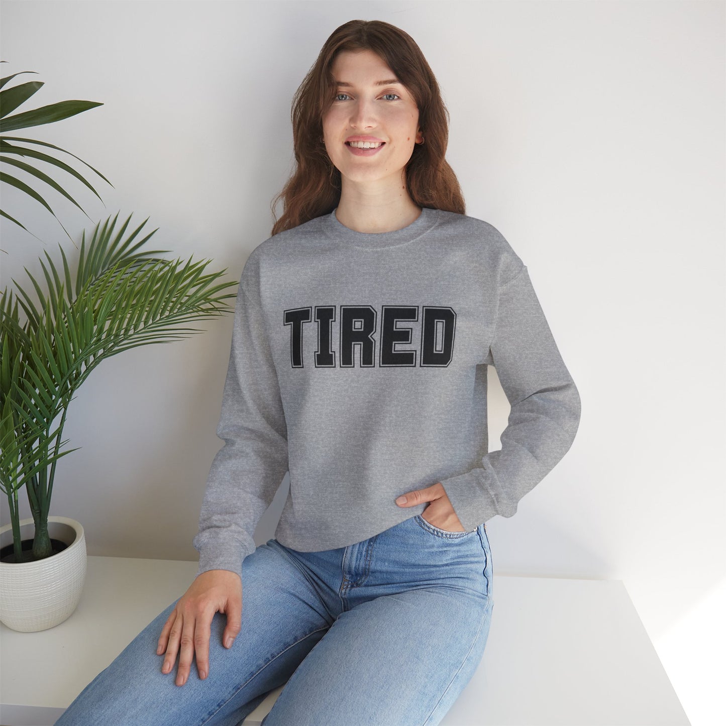 Model wearing grey “Tired” crewneck sitting on couch
